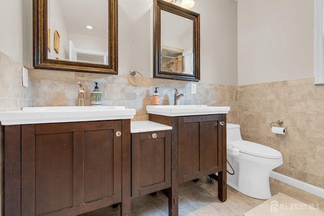 bathroom featuring toilet, a sink, a wainscoted wall, two vanities, and tile walls