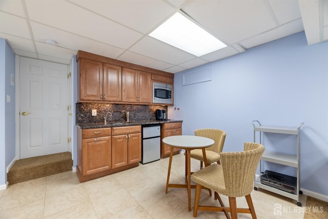 kitchen featuring brown cabinetry, dark stone counters, stainless steel microwave, backsplash, and a sink