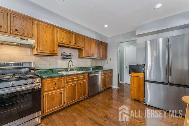 kitchen with dark countertops, a sink, under cabinet range hood, stainless steel appliances, and dark wood-style flooring