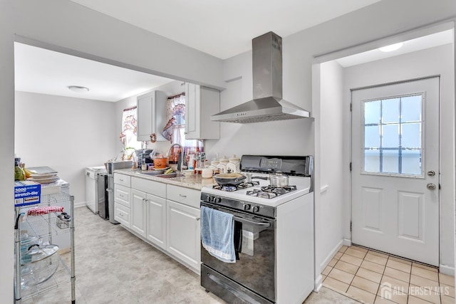 kitchen with white range with gas stovetop, wall chimney range hood, sink, light tile patterned floors, and white cabinetry