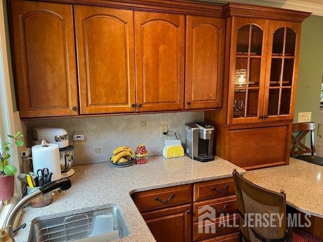 kitchen featuring a sink, backsplash, light stone countertops, brown cabinetry, and glass insert cabinets