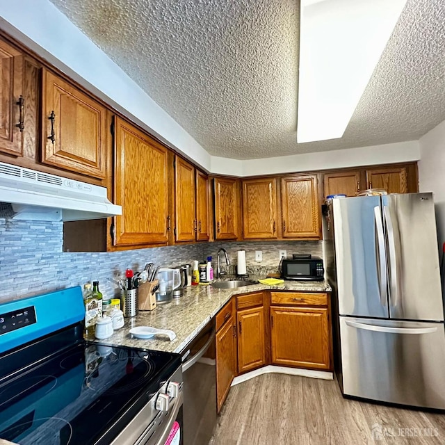 kitchen featuring under cabinet range hood, light wood-type flooring, appliances with stainless steel finishes, brown cabinetry, and a sink