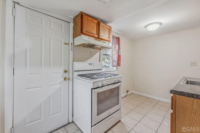 kitchen with baseboards, white gas range oven, light tile patterned floors, under cabinet range hood, and brown cabinetry