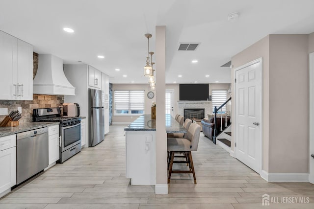 103 Rumford Way, Manalapan, NJ 07726 - kitchen with custom range hood, stainless steel appliances, light wood finished floors, a breakfast bar area, and dark stone countertops