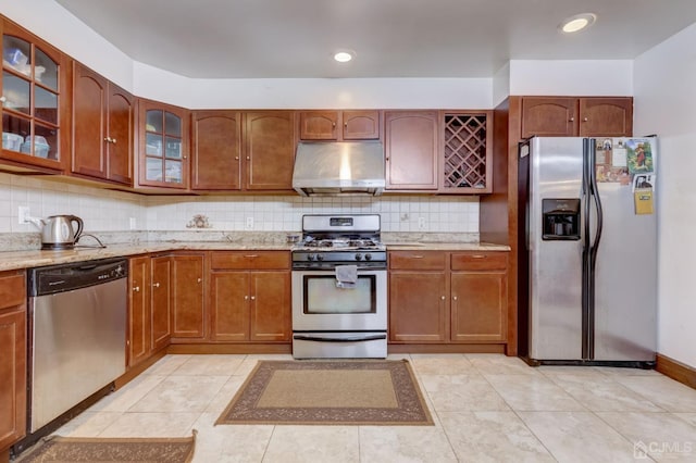 kitchen with light stone counters, decorative backsplash, glass insert cabinets, under cabinet range hood, and appliances with stainless steel finishes