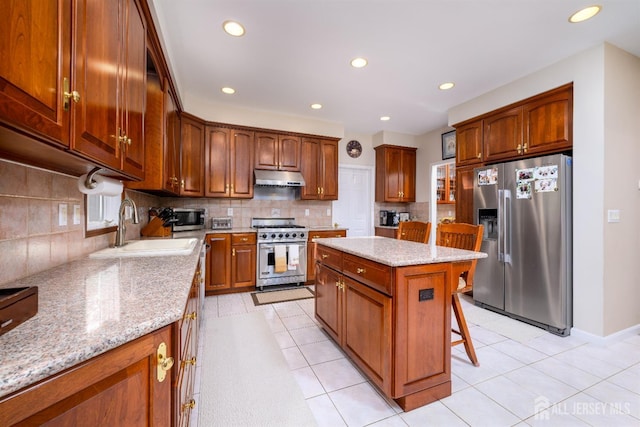 kitchen featuring under cabinet range hood, stainless steel appliances, a kitchen island, a sink, and light stone countertops