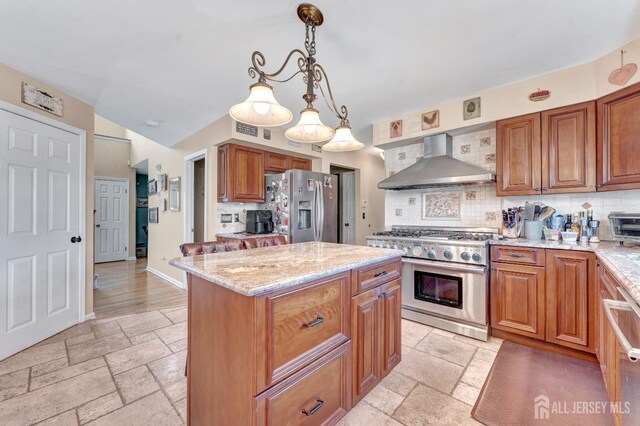 kitchen featuring stainless steel appliances, light stone counters, a kitchen island, decorative light fixtures, and wall chimney exhaust hood