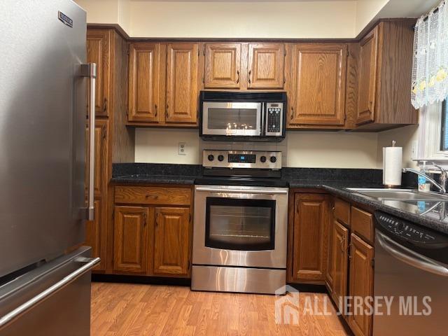 kitchen featuring brown cabinetry, dark countertops, a sink, light wood-style flooring, and stainless steel appliances
