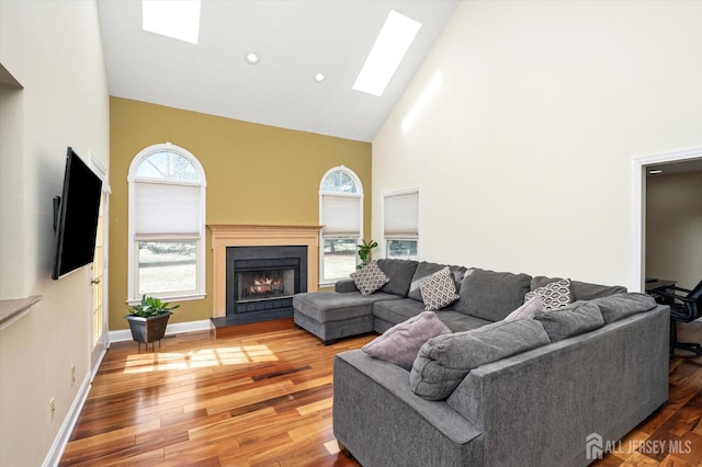 living area with baseboards, light wood-style flooring, a warm lit fireplace, a skylight, and high vaulted ceiling