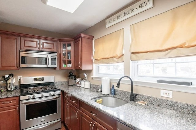 kitchen featuring light stone counters, a skylight, a sink, appliances with stainless steel finishes, and glass insert cabinets