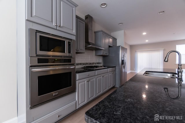 kitchen featuring decorative backsplash, wall chimney range hood, gray cabinets, a sink, and stainless steel appliances