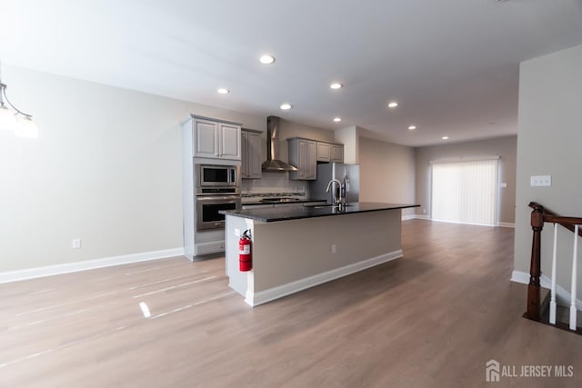 kitchen with wall chimney range hood, dark countertops, an island with sink, stainless steel appliances, and recessed lighting