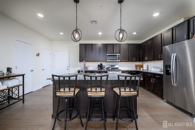 kitchen featuring dark brown cabinetry, tasteful backsplash, a center island with sink, appliances with stainless steel finishes, and pendant lighting