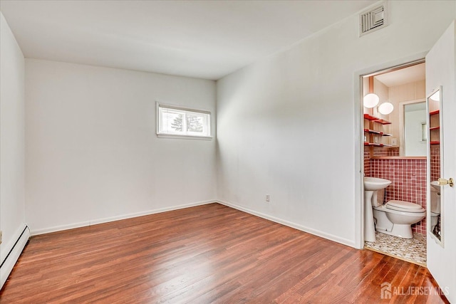 unfurnished bedroom featuring a baseboard radiator, wood finished floors, tile walls, and connected bathroom