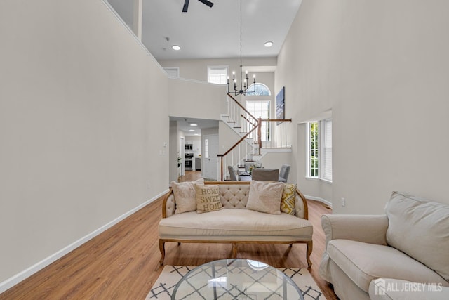 living area featuring baseboards, light wood finished floors, stairs, a towering ceiling, and ceiling fan with notable chandelier