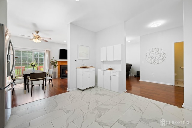 kitchen with ceiling fan, stainless steel fridge with ice dispenser, baseboards, a warm lit fireplace, and white cabinetry