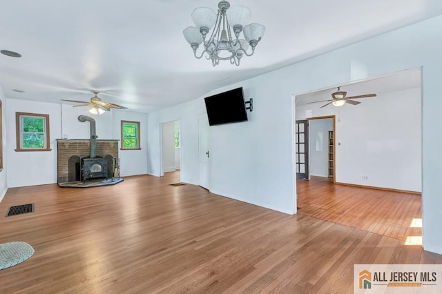 unfurnished living room with a wood stove, a wealth of natural light, wood-type flooring, and ceiling fan with notable chandelier