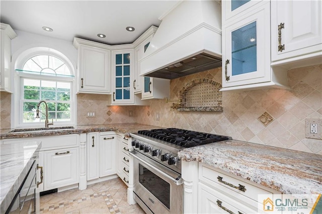 kitchen featuring light stone counters, premium range hood, a sink, white cabinetry, and high end range