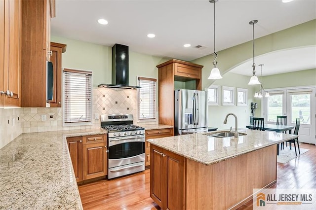 kitchen with wall chimney exhaust hood, sink, hanging light fixtures, stainless steel appliances, and a kitchen island with sink