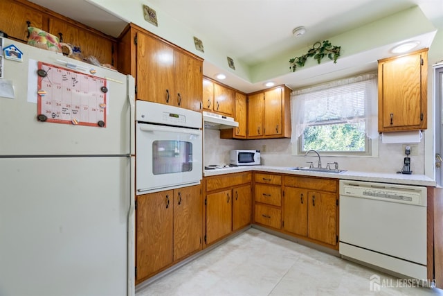 kitchen featuring brown cabinets, a sink, light countertops, under cabinet range hood, and white appliances