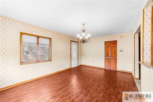 empty room featuring dark wood-style flooring, a chandelier, wallpapered walls, and baseboards