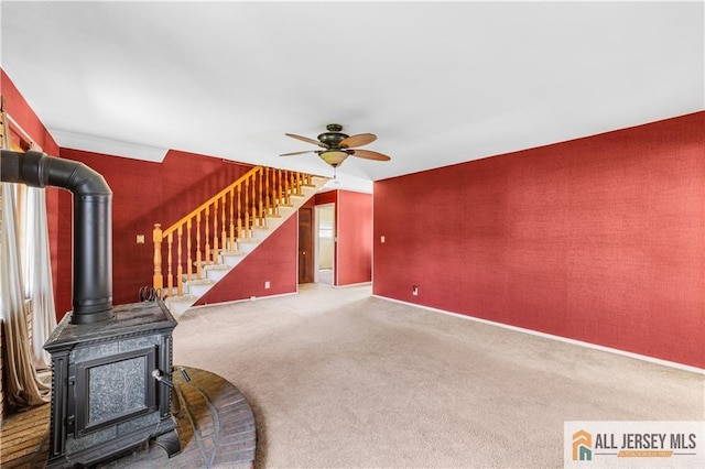 unfurnished living room featuring a wood stove, ceiling fan, carpet flooring, stairs, and baseboards