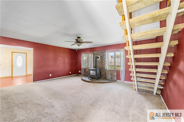 unfurnished living room with a wood stove, carpet floors, ceiling fan, and baseboards
