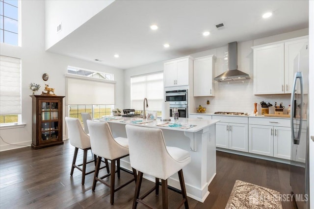 kitchen with wall chimney exhaust hood, stainless steel appliances, an island with sink, and white cabinets