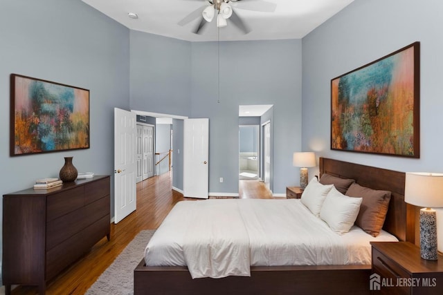 bedroom featuring a towering ceiling, a ceiling fan, ensuite bath, wood finished floors, and baseboards