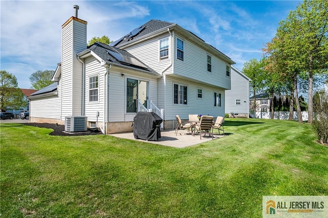 rear view of property with roof mounted solar panels, a chimney, a patio area, central air condition unit, and a lawn