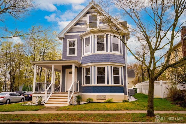 victorian-style house with a porch, central AC, fence, and a front lawn