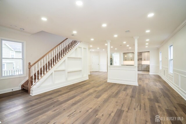 unfurnished living room featuring ornate columns, ornamental molding, wood finished floors, and stairway