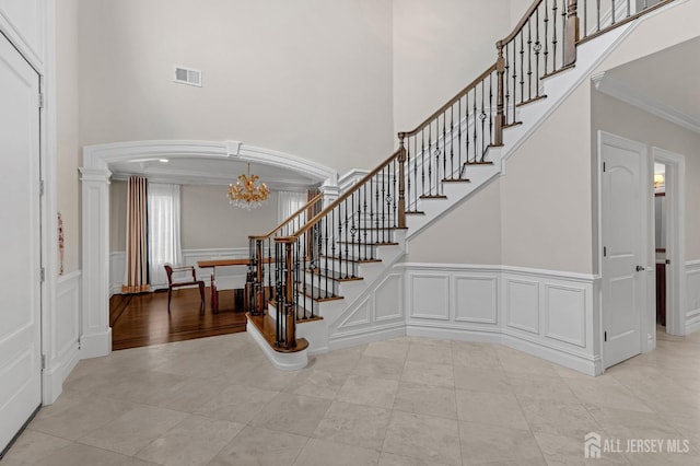 stairs featuring an inviting chandelier, crown molding, visible vents, a towering ceiling, and a decorative wall