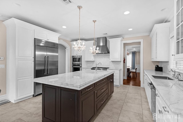 kitchen featuring white cabinets, a sink, built in appliances, wall chimney exhaust hood, and decorative backsplash