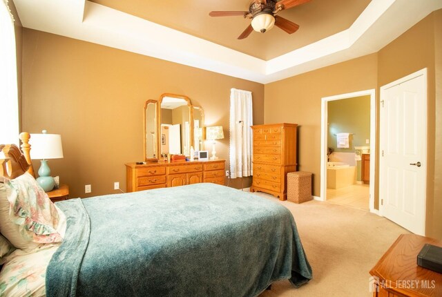 bedroom with a raised ceiling, light carpet, ensuite bath, and a ceiling fan