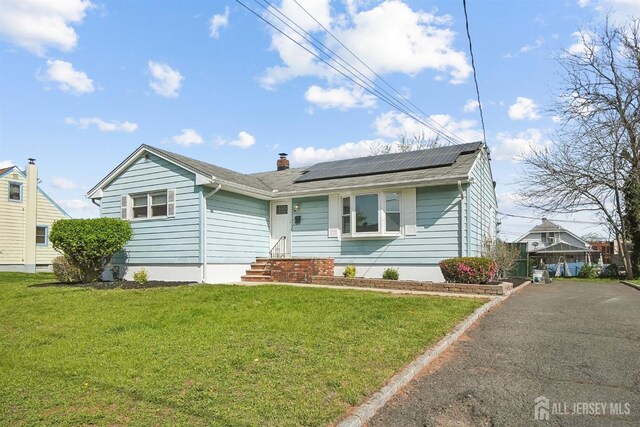single story home with aphalt driveway, a front lawn, roof mounted solar panels, and a chimney