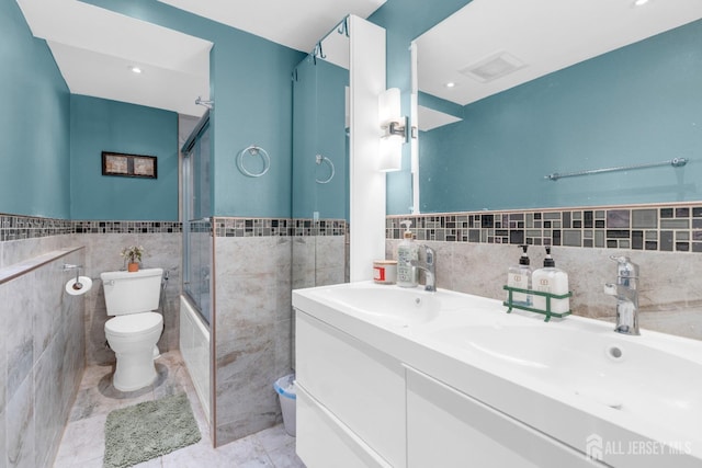 bathroom featuring tile walls, toilet, a wainscoted wall, bath / shower combo with glass door, and double vanity