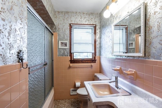bathroom with tile walls, toilet, wallpapered walls, a wainscoted wall, and combined bath / shower with glass door
