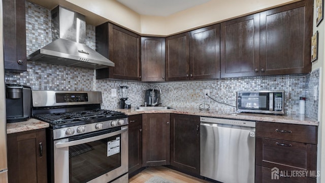 kitchen featuring decorative backsplash, appliances with stainless steel finishes, dark brown cabinetry, a sink, and wall chimney exhaust hood