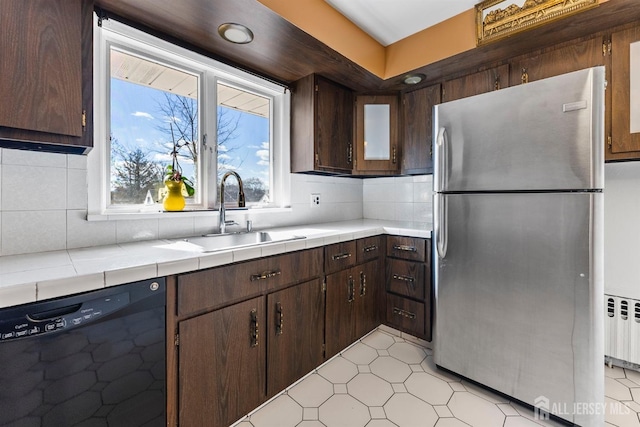 kitchen with a sink, black dishwasher, radiator heating unit, freestanding refrigerator, and tile counters