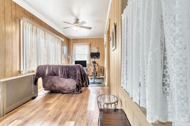 living area with radiator, wood finished floors, ceiling fan, wood walls, and crown molding