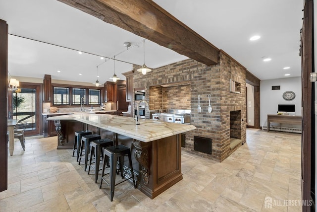 kitchen with a brick fireplace, beamed ceiling, a breakfast bar area, stone tile floors, and a large island with sink