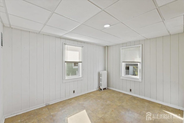 empty room featuring baseboards, a drop ceiling, radiator heating unit, and wood walls