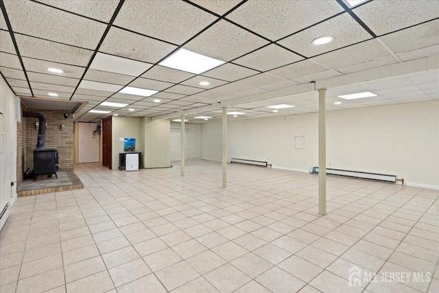 basement with a paneled ceiling, light tile patterned floors, a wood stove, and a baseboard heating unit