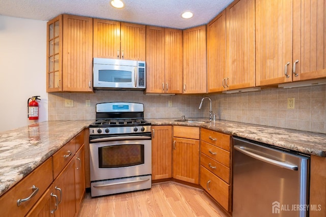 kitchen with brown cabinetry, light stone countertops, stainless steel appliances, light wood-type flooring, and a sink