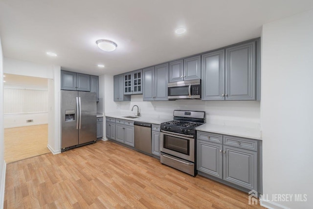 kitchen featuring appliances with stainless steel finishes, gray cabinetry, light wood finished floors, light countertops, and glass insert cabinets