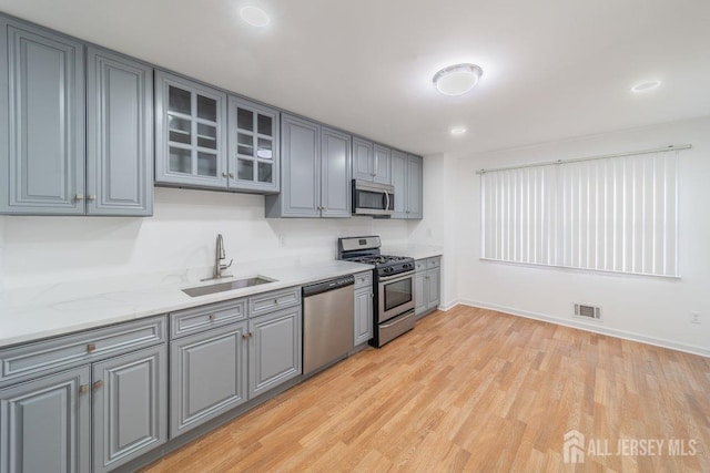 kitchen with appliances with stainless steel finishes, a sink, gray cabinets, light wood-style floors, and glass insert cabinets