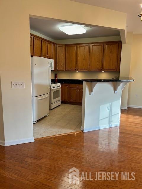 kitchen with white appliances, light wood finished floors, brown cabinetry, dark countertops, and a kitchen breakfast bar