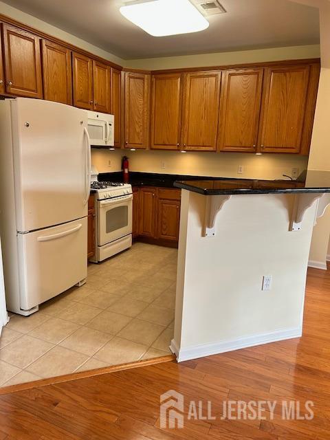 kitchen with a kitchen breakfast bar, dark countertops, brown cabinets, light wood-style floors, and white appliances