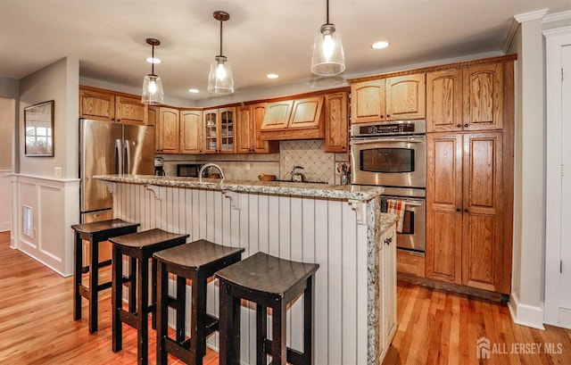 kitchen featuring brown cabinetry, light wood finished floors, stainless steel appliances, glass insert cabinets, and backsplash